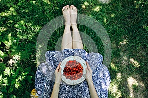 Barefoot girl is sitting on the grass in blue vintage dress. Woman is holding plate with home strawberries. Rustic summer fruit