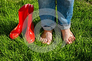 Barefoot girl standing next red garden gumboots on grass