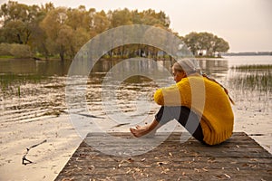 A barefoot girl in a yellow knitted sweater sits on the bridge over the lake