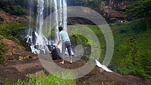 Barefoot guy with backpack climbs up stone by waterfall