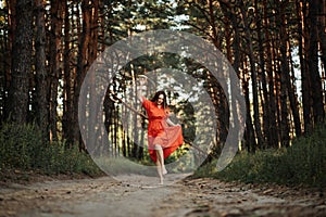 Barefoot happy young woman in red dress with hand raised dancing in pine forest at summer day.