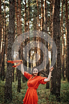 Barefoot happy young woman in red dress with hand raised dancing in pine forest at summer day.