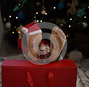 barefoot kid lies in front of the Christmas tree, the numbers 2023 are written on his feet, a souvenir of Santa