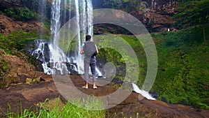 Barefoot man with backpack climbs up rock at waterfall