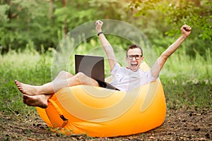 Barefoot man with hands up in winner pose and happy screaming, in shirt, red bow tie and glasses lying on inflatable sofa with