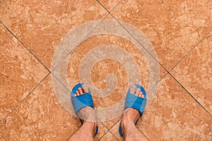 Barefoot men's feet in blue home slippers stand on a brown or orange floor tile ceramic background, view from above