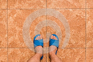 Barefoot men's feet in blue home slippers stand on a brown or orange floor tile ceramic background, view from above
