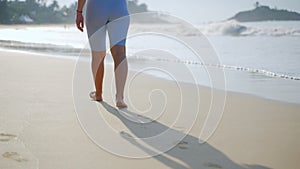 Barefoot person in blue shorts strolls on sandy shore, waves lap gently, tranquil coastal walk. Footprints in sand