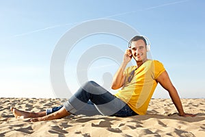 Barefoot smiling man sitting in sand at the beach listening to music with headphones