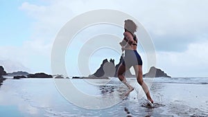 Barefoot sporty girl with slim body running along sea surf by water pool to keep fit and health. Beach background with
