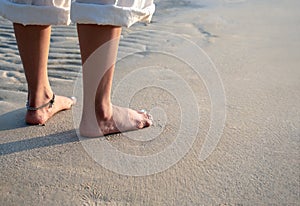 Barefoot woman in white pants with folded legs standing on quiet sandy beach in the warm sunlight.