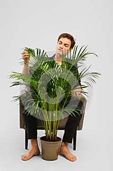 Barefoot young man in black suit siting in an armchair and looking at home plant