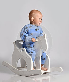 Barefooted baby boy in blue fleece jumpsuit with stars plays has fun riding white kids rocking horse toy swinging