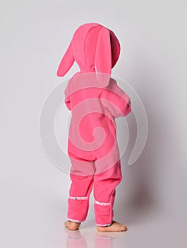 Barefooted baby girl in pink warm comfortable jumpsuit with hood on and bunny ears stands back to camera