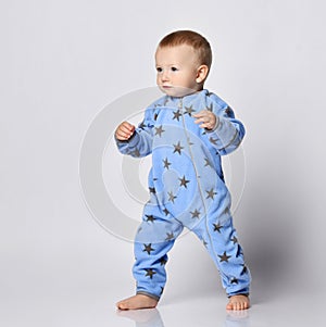 Barefooted blond baby boy toddler in blue fleece jumpsuit with stars stands steadily with his legs spread wide.