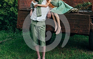 A barefooted girl in a green dress sits on the back of a car and hugs a barefoot, next to a standing guy, a summer day in the vill