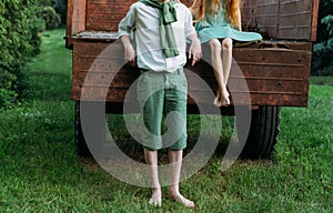A barefooted girl in a green dress is sitting in an old caravan, and beside her, barefoot, on the grass, stands a guy in a white s