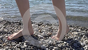 Barefooted woman walking on pebble sea beach near water, legs close-up.