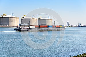 Barge loaded with containers sailing past large concrete tanks for liquefied natural gas storage