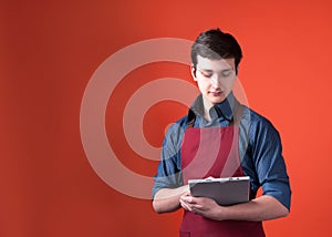 Barista with dark hair in burgundy apron holding and looking at digital tablet on orange background
