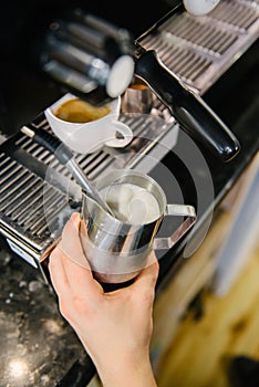 Barista froths milk for coffee. There is a cup of freshly brewed coffee in the coffee machine photo