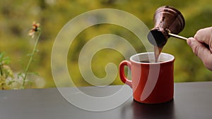 Barista hand pouring tasty hot coffee in red cup brewed in traditional turkish pot, cafe table outdoors