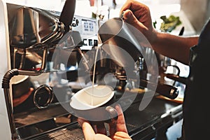 Barista man pouring whipped milk from frothing pitcher in cup with coffee standing near professional coffee machine in