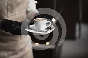 Barista wearing black medical gloves carries a cup of espresso in a coffee shop. Selective focus. Protecting health