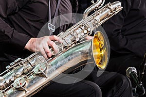 The baritone saxophone lies on the knee of the musician in a black shirt and trousers. The right hand lies on a wooden wind instru