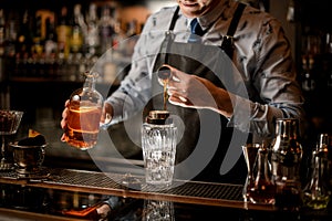 Barman in black apron pours drink to glassy shaker using beaker.