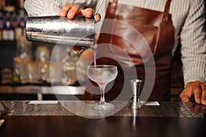Barman in a brown apron pours from a steel shaker alcohol cocktail