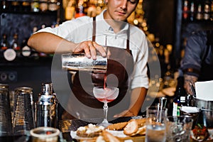 Barman in a brown leather apron pouring fruit alcoholic cocktail into the glass