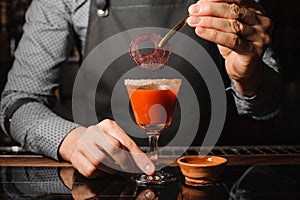 Barman decorating a cocktail glass filled with red alcoholic drink with salt and beet