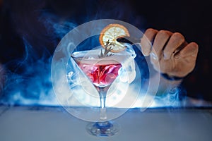 Barman make red alcoholic cocktail with smoke in glass, decorated with orange