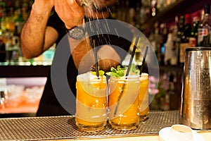 Barman making orange hurricane cocktail indoors of nightclub on top pouring sugar over green mint leaves