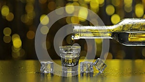 Barman pour frozen vodka from bottle into shot glass. Ice cubes against shiny gold party background