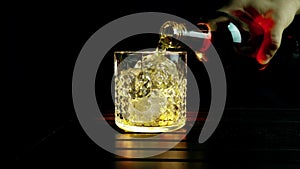 Barman pouring whiskey in the glass with ice cubes on wood table and black dark background, focus on ice cubes, whisky relax time