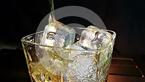 Barman pouring whiskey in the glass with ice cubes on wood table and black dark background, focus on ice cubes, whisky relax time