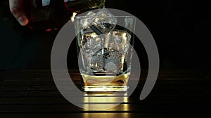 Barman pouring whiskey in the glass with ice cubes on wood table and black dark background, focus on ice cubes, whisky relax time