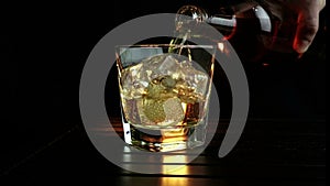 Barman pouring whiskey in the glass with ice cubes on wood table and black dark background, focus on ice cubes, whisky relax time