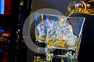Barman pouring whiskey in two glasses with ice cubes on table with light tint blue and reflection