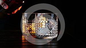 Barman pouring whiskey in the two glasses with ice cubes on wood table and black dark background, focus on ice cubes