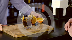 Barman puts alcohol cocktail on the rocks glass to client in bar counter. Hands close-up.