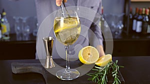 Barman puts lemon and rosemary in alcohol cocktail with sparkling wine, ice and liquor in wineglass, close-up view.