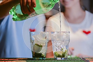 Barmans hand with soda bottle pouring cocktail into two glasses arranged in a line. Two bartenders at work in bar.