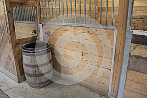 A barrel outside a stall in a stable with wooden plank walls and sliding doors with metal bars