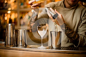 Bartender carefully pouring drink from metal jigger into mixing cup on bar counter