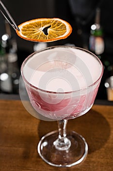 Bartender is decorating pink Clover club alcoholic cocktail with orange slice at the bar. Bartender mixes egg white