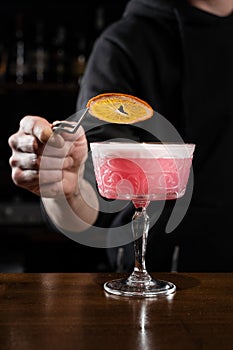 Bartender is decorating pink Clover club alcoholic cocktail with orange slice at the bar. Bartender mixes egg white
