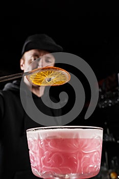 Bartender is decorating pink Clover club alcoholic cocktail with orange slice at the bar. Bartender mixes egg white